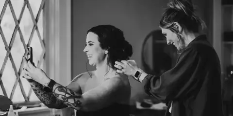 A black and white photo of a hairstylist styling a bride's hair while the bride takes a selfie.