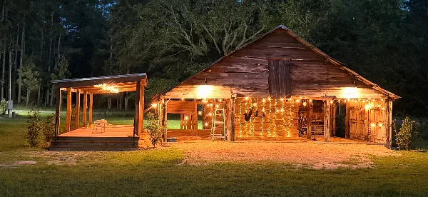 A rustic wooden barn and covered outdoor event area illuminated by string lights at dusk, set against a forest background.