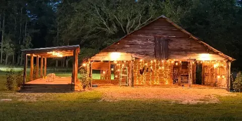 A rustic wooden barn and covered outdoor event area illuminated by string lights at dusk, set against a forest background.