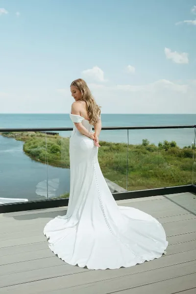 A bride in a white gown standing on a wooden balcony overlooking a lake and shoreline.