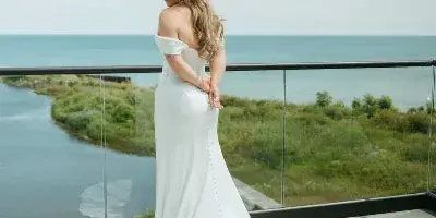 A bride in a white gown standing on a wooden balcony overlooking a lake and shoreline.