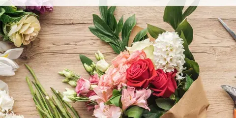 A hand holding a mixed flower bouquet with red roses, pink alstroemeria, and white blooms wrapped in brown paper.