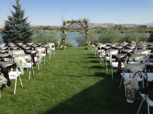 Outdoor wedding ceremony setup with white chairs and floral arch overlooking a river and hills.