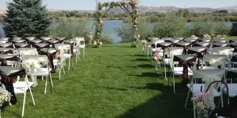 Outdoor wedding ceremony setup with white chairs and floral arch overlooking a river and hills.
