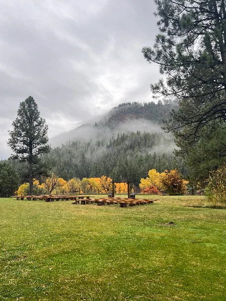 A wooden wedding ceremony arch and seating area on a grassy field with a misty mountain in the background.