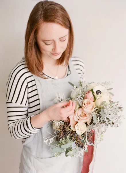 A woman in a striped shirt and apron arranging a wedding bouquet with roses and greenery.