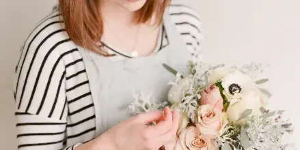 A woman in a striped shirt and apron arranging a wedding bouquet with roses and greenery.