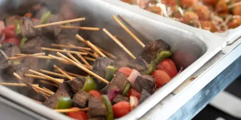 A close-up of a catering buffet featuring skewers of meat and vegetables in a stainless steel serving tray.