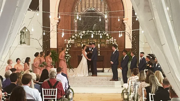A wedding ceremony in progress inside a church sanctuary with a bride and groom at the altar, surrounded by wedding party members and seated guests.
