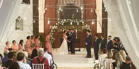 A wedding ceremony in progress inside a church sanctuary with a bride and groom at the altar, surrounded by wedding party members and seated guests.