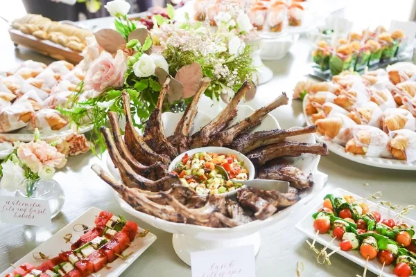 A catering table display featuring a central platter of grilled lamb ribs with a vegetable relish, surrounded by watermelon and feta skewers, caprese skewers, crostini, and floral centerpieces.