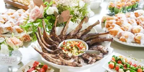 A catering table display featuring a central platter of grilled lamb ribs with a vegetable relish, surrounded by watermelon and feta skewers, caprese skewers, crostini, and floral centerpieces.