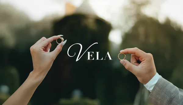 Two hands holding wedding rings against a blurred outdoor background with the text 'Vela' overlaid.