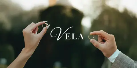 Two hands holding wedding rings against a blurred outdoor background with the text 'Vela' overlaid.