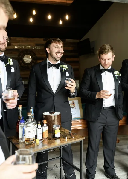 Groom and groomsmen in black tuxedos standing in a rustic venue interior with a wooden wall and a bar table.