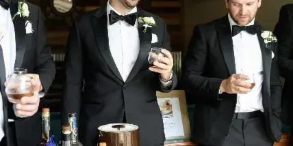 Groom and groomsmen in black tuxedos standing in a rustic venue interior with a wooden wall and a bar table.