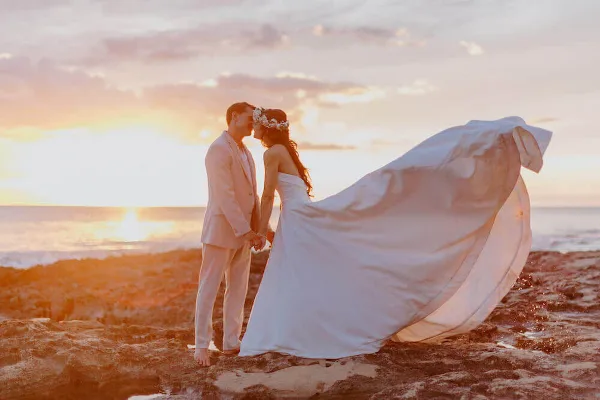 A bride and groom standing on a rocky shoreline at sunset, with the bride's dress blowing in the wind.