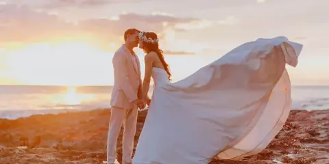 A bride and groom standing on a rocky shoreline at sunset, with the bride's dress blowing in the wind.