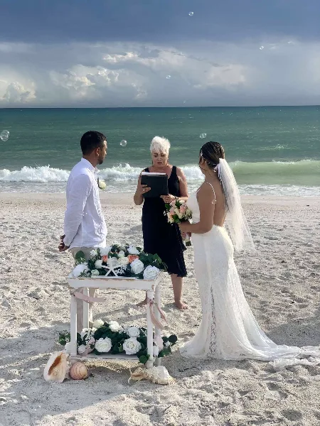A wedding officiant performing a ceremony for a couple on a sandy beach with the ocean in the background.