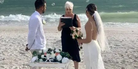 A wedding officiant performing a ceremony for a couple on a sandy beach with the ocean in the background.