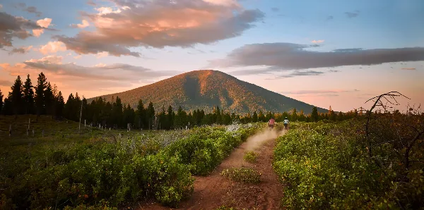 A scenic landscape view of a mountain at sunset with a dirt trail and cyclists in the foreground.