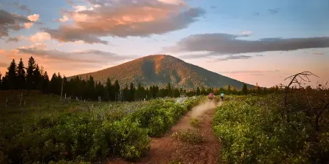 A scenic landscape view of a mountain at sunset with a dirt trail and cyclists in the foreground.