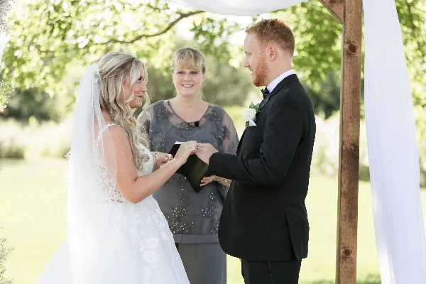 A bride and groom exchanging rings during an outdoor wedding ceremony with an officiant standing between them.