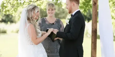 A bride and groom exchanging rings during an outdoor wedding ceremony with an officiant standing between them.