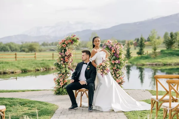 A bride and groom posing for a wedding portrait outdoors near a pond with a floral arch and mountains in the background.