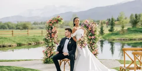 A bride and groom posing for a wedding portrait outdoors near a pond with a floral arch and mountains in the background.