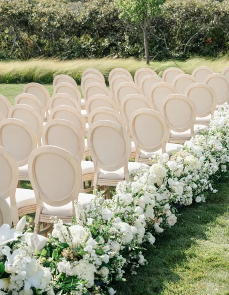 Rows of cream-colored upholstered chairs set up for an outdoor wedding ceremony with white floral arrangements.