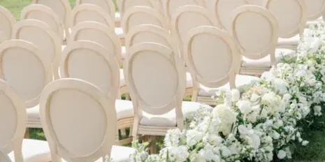 Rows of cream-colored upholstered chairs set up for an outdoor wedding ceremony with white floral arrangements.