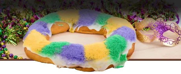 A King Cake with white icing and purple, green, and gold sugar, displayed with Mardi Gras beads and a mask.