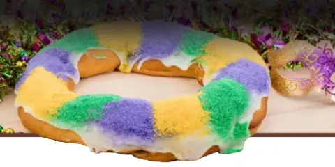 A King Cake with white icing and purple, green, and gold sugar, displayed with Mardi Gras beads and a mask.