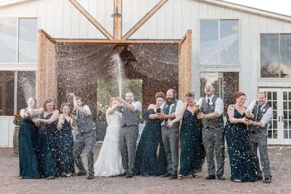 A wedding party spraying champagne in front of a rustic white barn with large wooden doors.