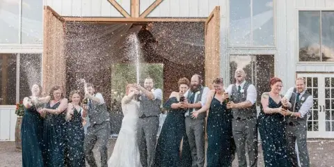 A wedding party spraying champagne in front of a rustic white barn with large wooden doors.