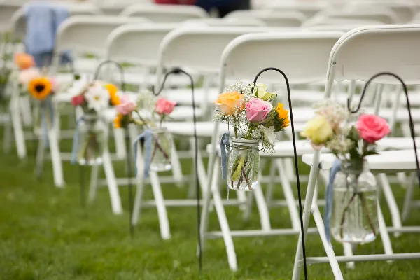 Rows of white folding chairs decorated with floral arrangements in mason jars at an outdoor wedding venue.