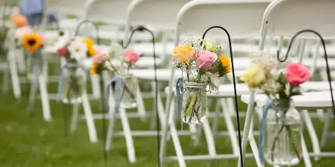 Rows of white folding chairs decorated with floral arrangements in mason jars at an outdoor wedding venue.