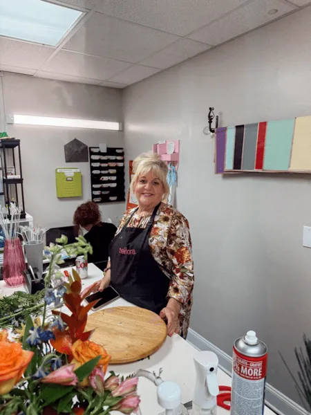 A female florist wearing an apron stands in a flower shop studio, arranging a bouquet of orange and pink flowers on a workbench.