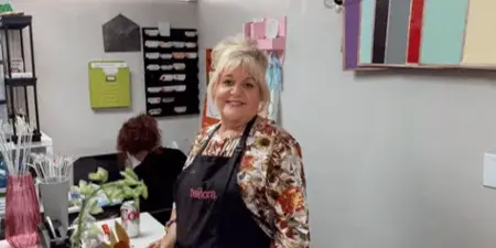 A female florist wearing an apron stands in a flower shop studio, arranging a bouquet of orange and pink flowers on a workbench.