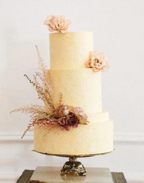 A three-tiered cream-colored wedding cake with textured piping and floral accents on a gold stand.
