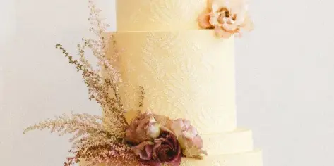 A three-tiered cream-colored wedding cake with textured piping and floral accents on a gold stand.
