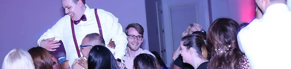 A DJ in a white shirt and suspenders interacting with a crowd of guests at a dance event.