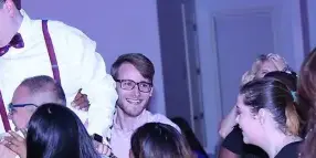 A DJ in a white shirt and suspenders interacting with a crowd of guests at a dance event.