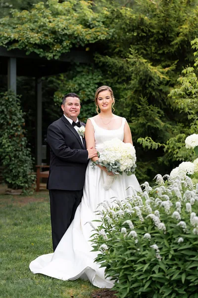 A bride in a white gown holding a bouquet of white flowers stands with a groom in a black tuxedo in a garden setting.