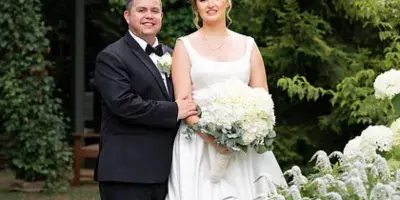 A bride in a white gown holding a bouquet of white flowers stands with a groom in a black tuxedo in a garden setting.