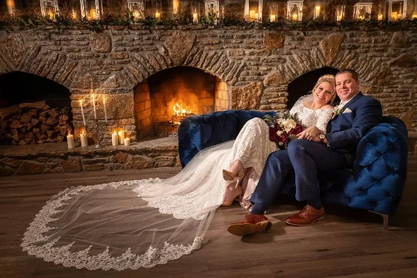 A bride and groom sitting on a blue sofa in front of a stone fireplace with candles and a fire.