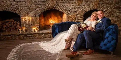 A bride and groom sitting on a blue sofa in front of a stone fireplace with candles and a fire.