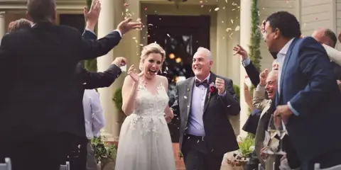 A bride and groom walking through a confetti shower with cheering guests at a wedding reception.