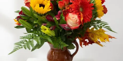 Mixed bouquet of sunflowers, roses, and lilies in a brown pitcher vase